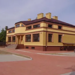 Dwukondygnacyjny budynek z cegły klinkierowej w odcieniach żółci i brązu, z brązowym dachem i czterema kominami, otoczony kostką brukową i zielenią.