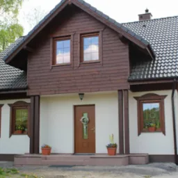 Gotowy dom z drewnianą elewacją i dekoracyjnymi oknami, zlokalizowany w spokojnej okolicy w Ostrowie Wielkopolskim. Widoczny wjazd do garażu i elementy zieleni.