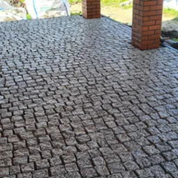 Świeżo ułożona, mokra kostka granitowa w odcieniach szarości tworzy wzór na podjeździe, widoczne ceglane filary i worki z materiałem budowlanym w tle.