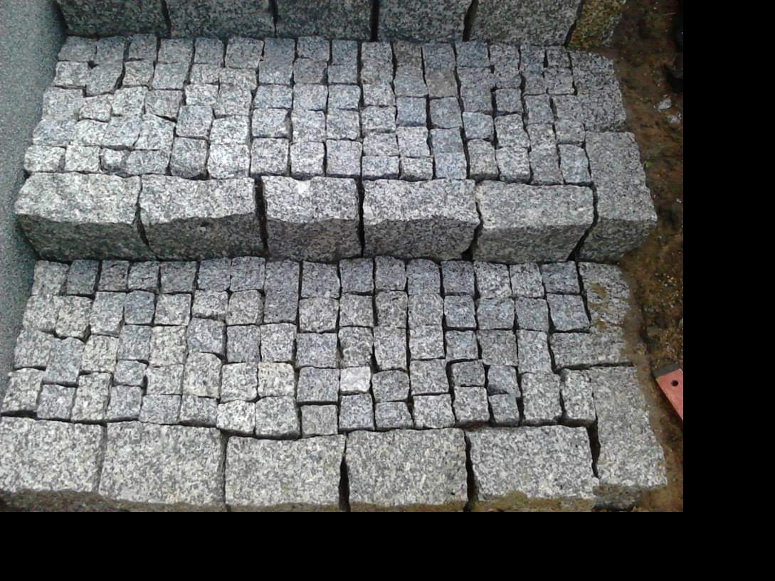 Stosy szarej kostki granitowej o różnych rozmiarach, przygotowane do ułożenia, z widocznym fragmentem poziomicy w prawym dolnym rogu.