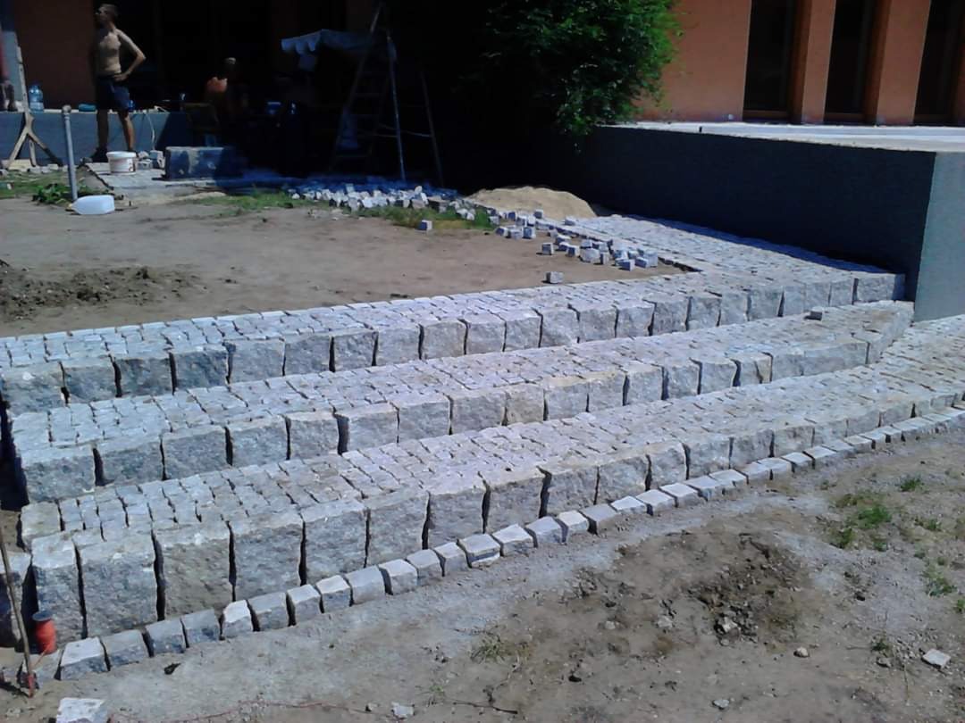 Schody z kostki granitowej w trakcie budowy, widoczne nierówności terenu i przygotowane materiały, budynek w tle.