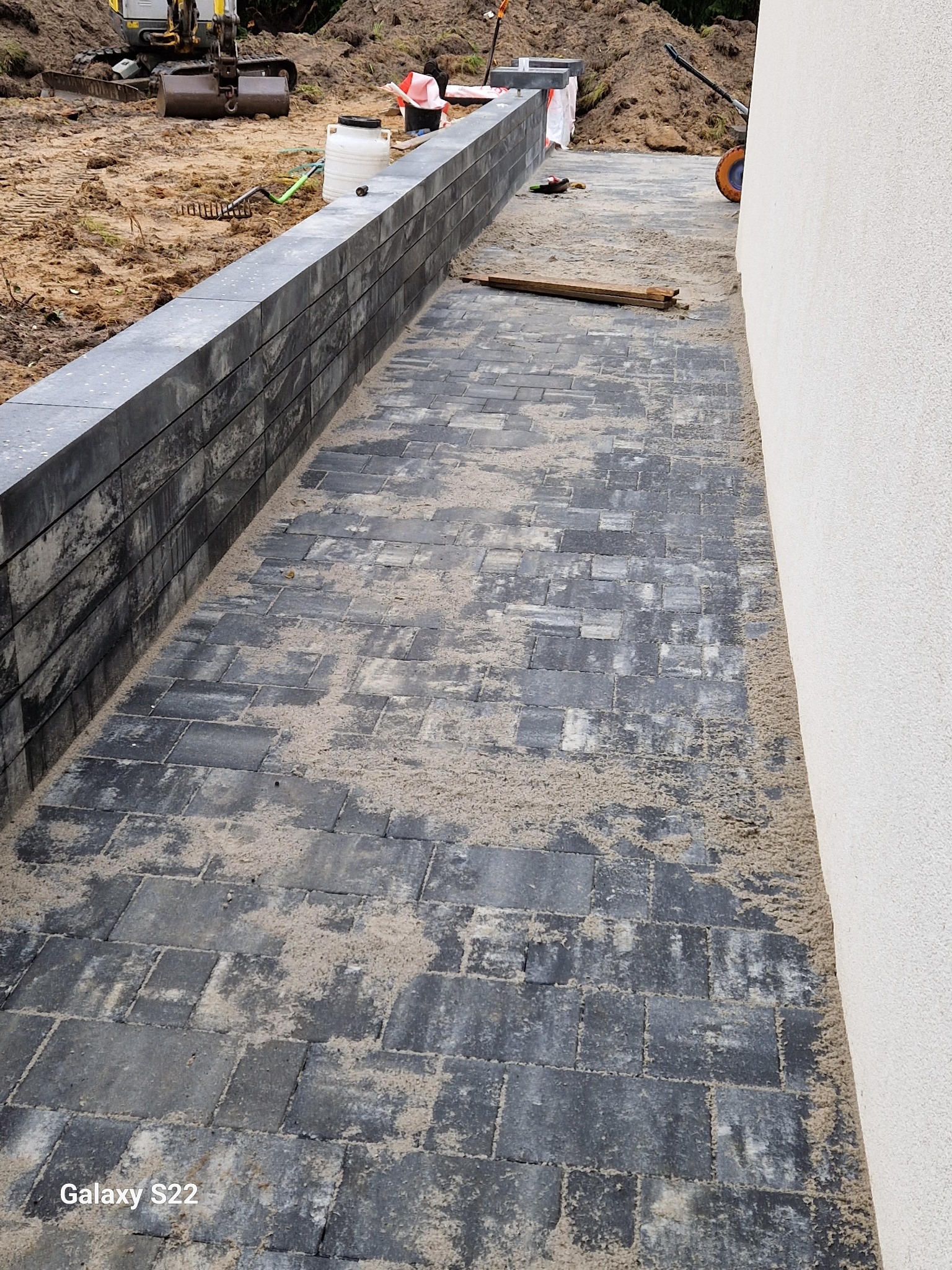 Ułożona, szara kostka brukowa zasypana piaskiem, widoczna ściana budynku, mur z szarych elementów, w tle sprzęt budowlany i ziemia.