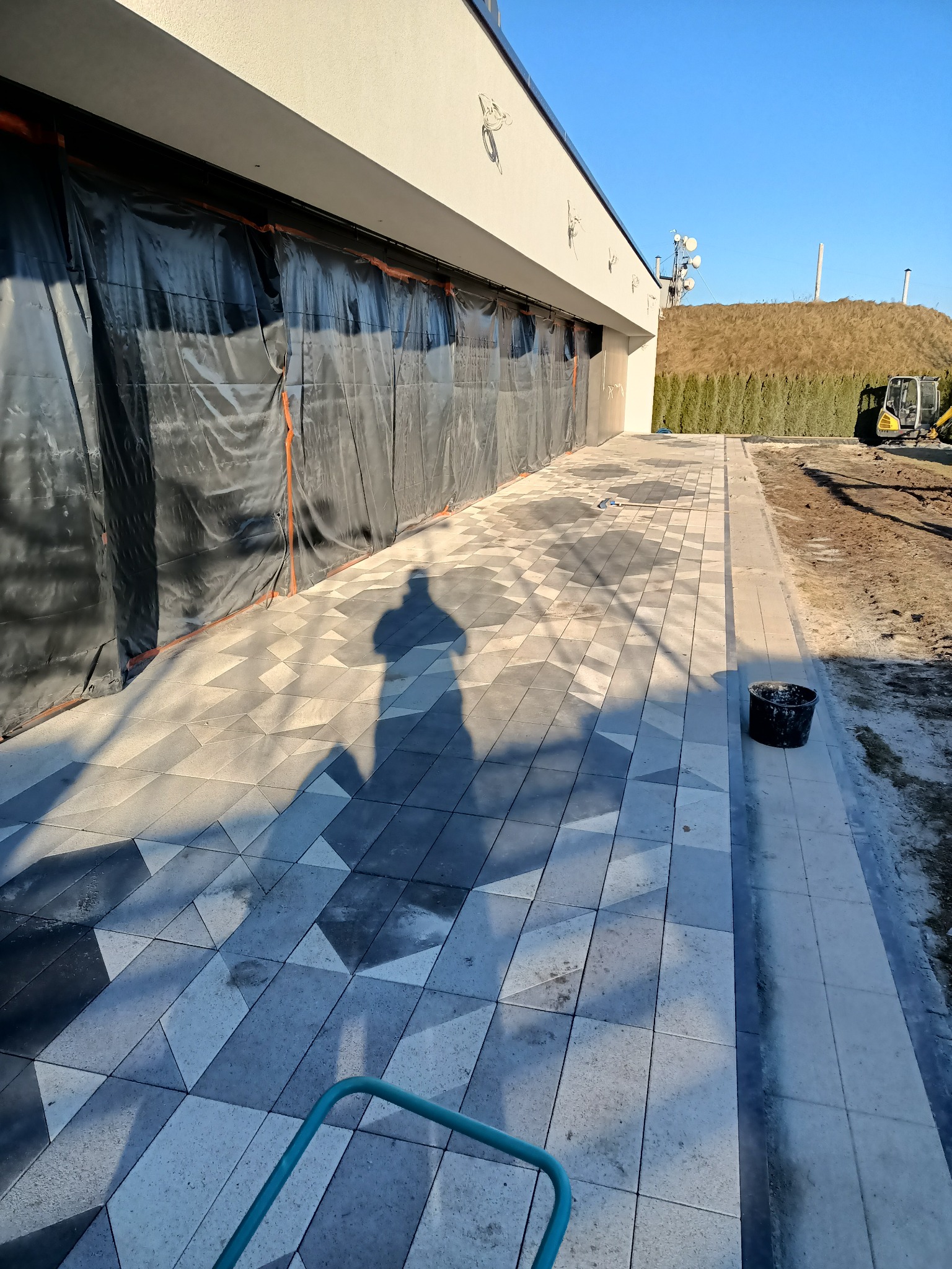Układanie kostki brukowej w geometryczny wzór na posesji, widoczny cień osoby fotografującej, zabezpieczona folią elewacja budynku w tle, teren budowy.