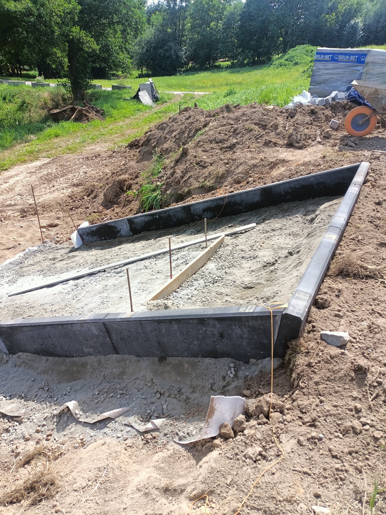 Przygotowany teren pod ułożenie kostki brukowej z obrzeżami, widoczne metalowe pręty, drewniane łaty, podsypka piaskowa i rolka żółtej linki, w tle trawnik i drzewa.