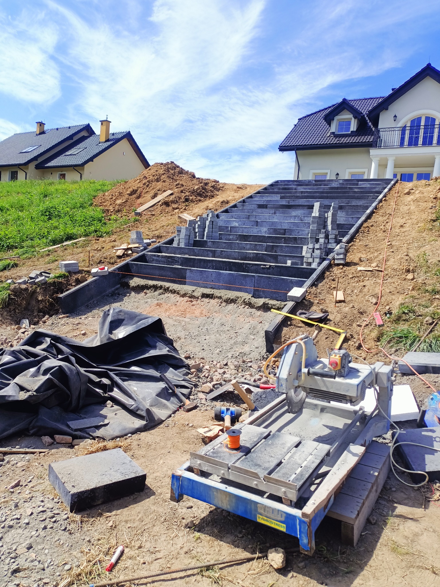 Budowa schodów zewnętrznych z kostki brukowej na wzgórzu przed domem, widoczna maszyna do cięcia kostki i przygotowane elementy.