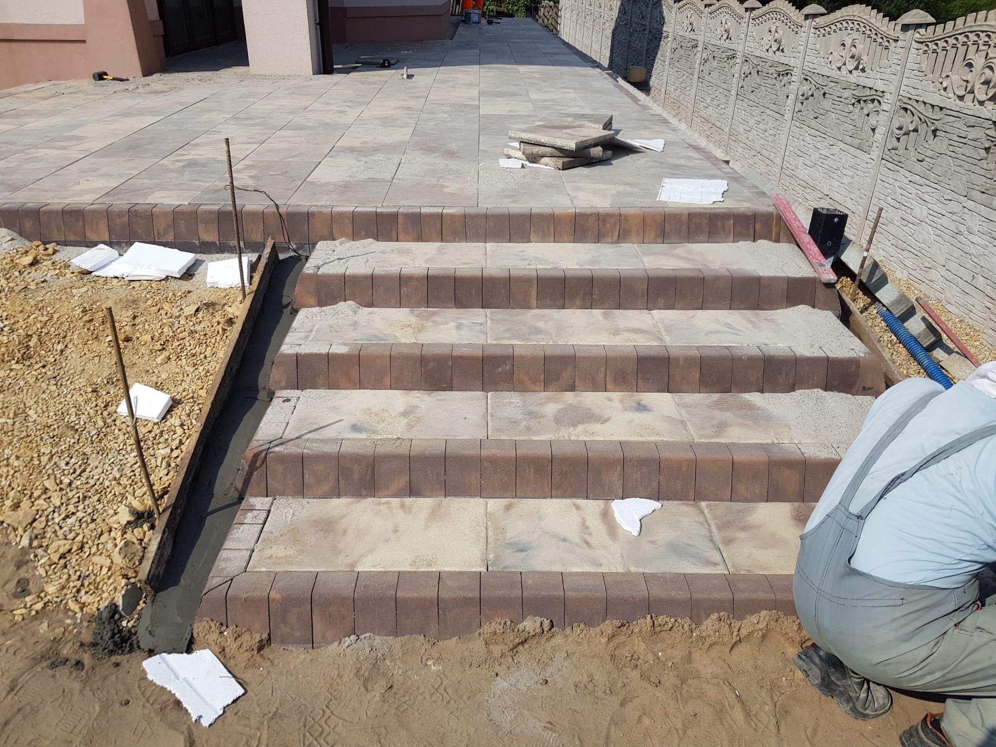 Budowa schodów z kostki brukowej z brązowym obrzeżem, widoczne etapy prac, piasek na stopniach, fragment chodnika i ogrodzenia w tle, w kadrze fragment sylwetki pracownika w kombinezonie.