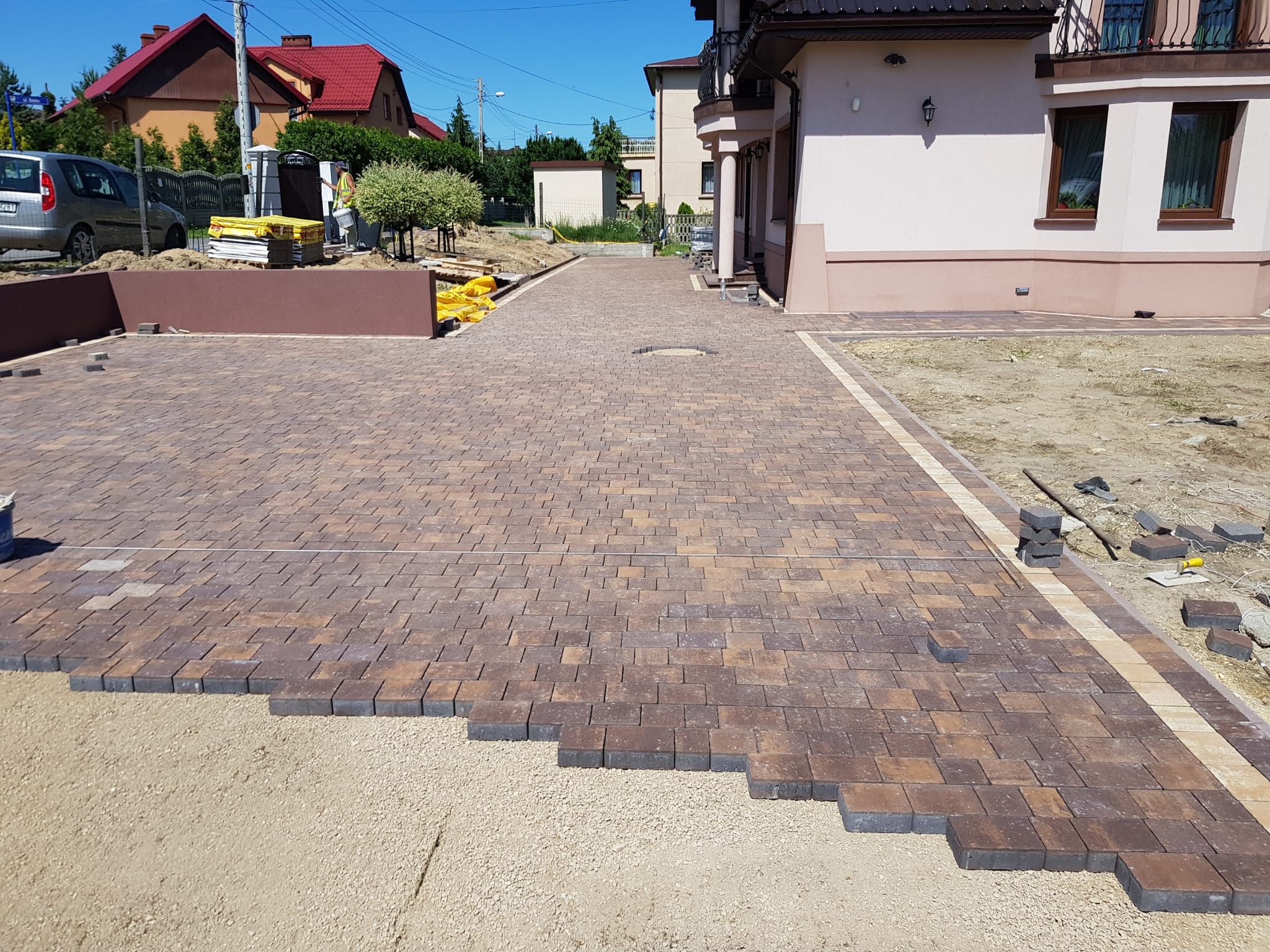 Brązowo-beżowa kostka brukowa w trakcie układania na podjeździe, widoczne przygotowane podłoże i obrzeża, dom w tle.