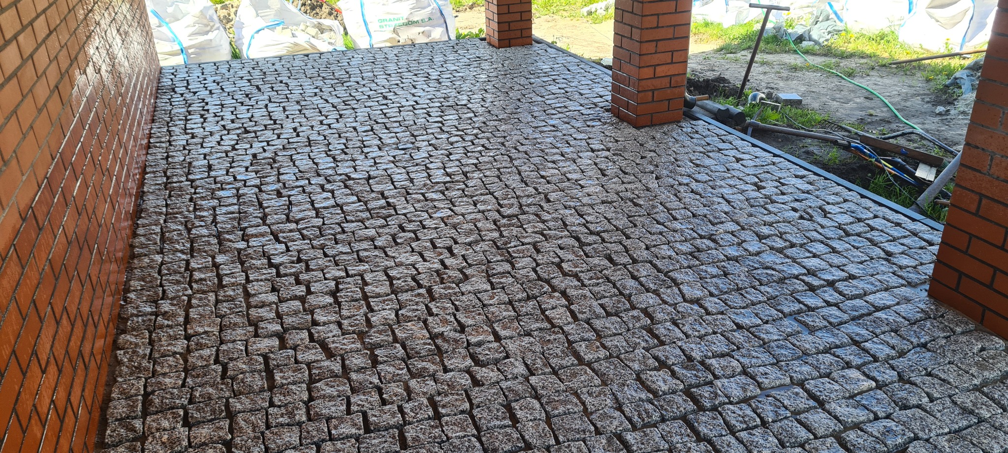 Świeżo ułożona, mokra kostka granitowa w odcieniach szarości tworzy wzór na podjeździe, widoczne ceglane filary i worki z materiałem budowlanym w tle.