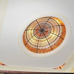 Ozdobny, okrągły witraż wpuszczony w biały sufit z delikatnymi sztukateriami, widziany z dołu pod kątem.