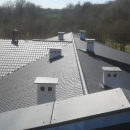Widok z góry na fragment dachu z dwoma rodzajami pokrycia: szara blachodachówka i czarna blacha trapezowa, z kominami wentylacyjnymi i pracującym dekarzem w oddali.