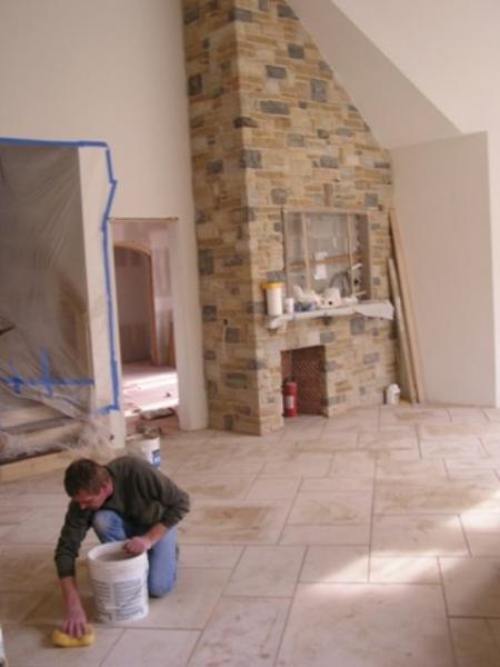 Mężczyzna czyści nowo położoną, jasną podłogę z płytek w przestronnym salonie z kamiennym kominkiem; widoczne wiadro i gąbka.