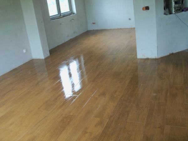 Błyszcząca, nowa podłoga imitująca drewno w surowym, jasnym wnętrzu. Widoczne refleksy światła na powierzchni. Ściany w stanie deweloperskim, widoczne ślady po pracach wykończeniowych.