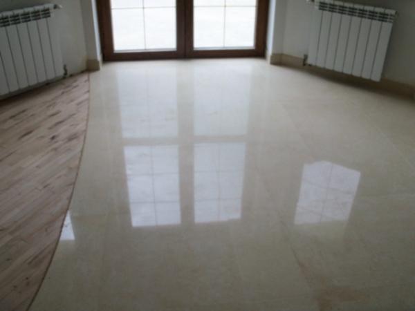 Błyszcząca, beżowa posadzka z płytek ceramicznych odbija światło z okna, widoczne dwa grzejniki i fragment drewnianej podłogi.