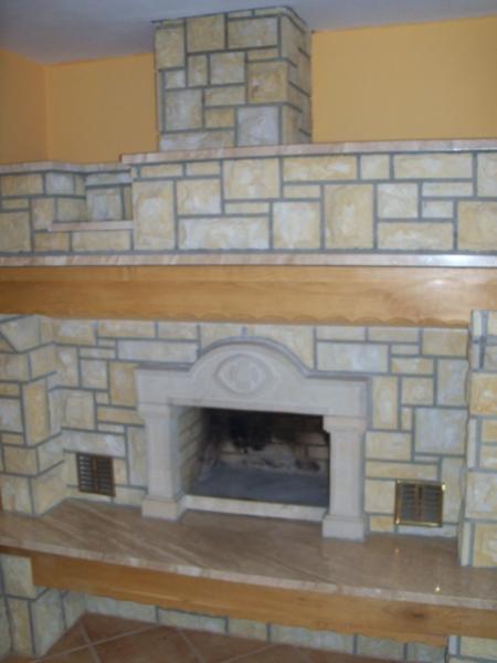 Obudowa kominka z piaskowca ułożonego w geometryczny wzór, z drewnianą półką i marmurowym blatem, w tle pomarańczowa ściana.