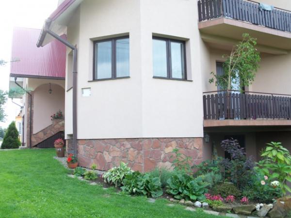 Dwupiętrowy dom z elewacją w kolorze kremowym i kamienną podmurówką w odcieniach brązu i czerwieni, z balkonem z drewnianą balustradą i zadbanym ogrodem z zielonym trawnikiem i rabatami kwiatowymi.