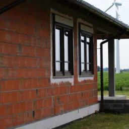 Dom w trakcie budowy z dwoma oknami z brązowymi ramami i częściowo zamontowanymi roletami zewnętrznymi, ceglana ściana, widoczny w tle wiatrak i zielone pole.
