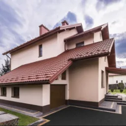 Dom z nowym dachem z dachówki ceramicznej w Koninie. Widoczny fragment ogrodu, błękitne niebo z chmurami i fragment wiaty garażowej.