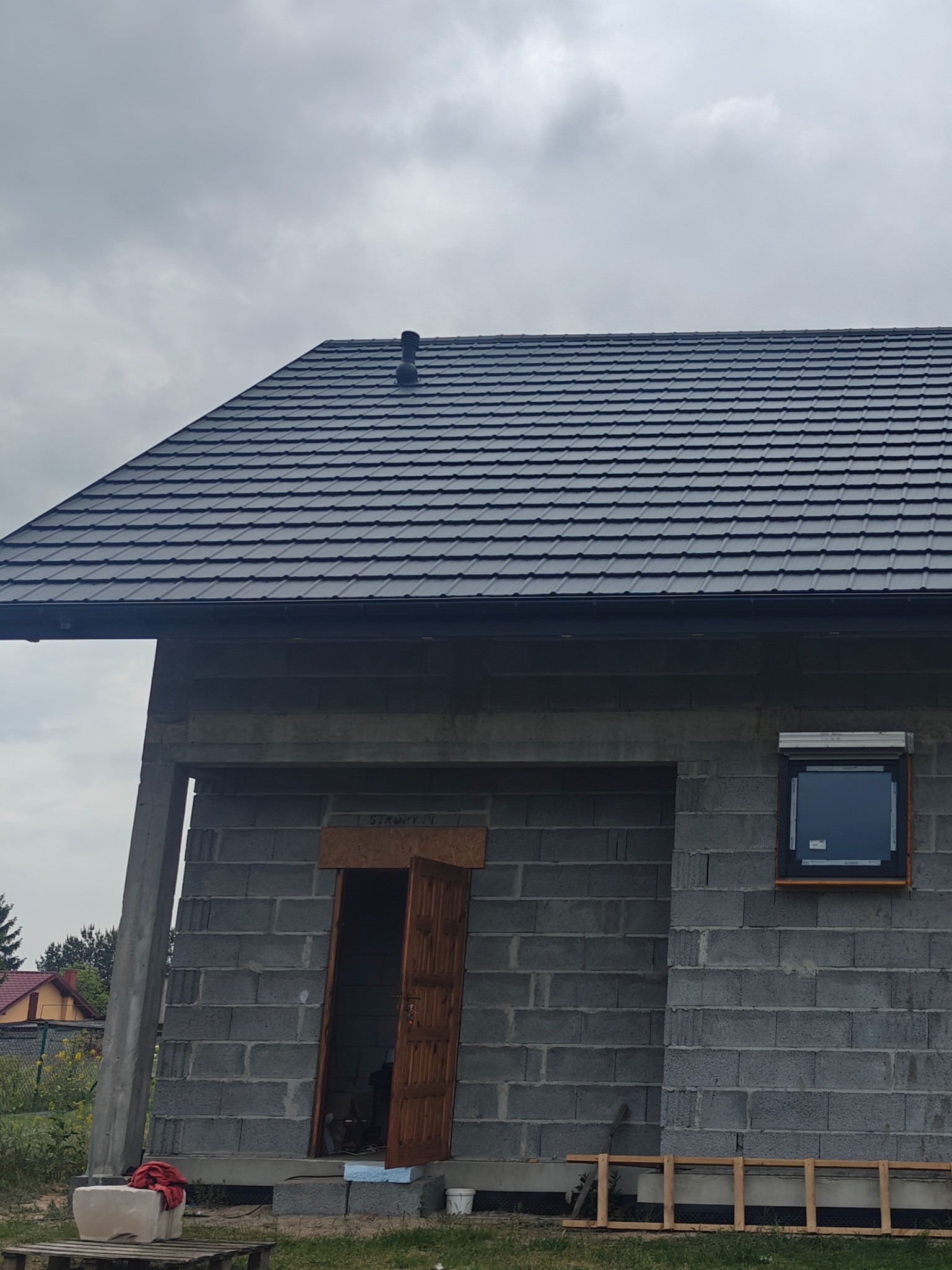 Nowoczesny dach z czarnej dachówki na surowym budynku z bloczków, widoczne okno z roletą, drabina i elementy budowlane na palecie. Pochmurne niebo w tle.