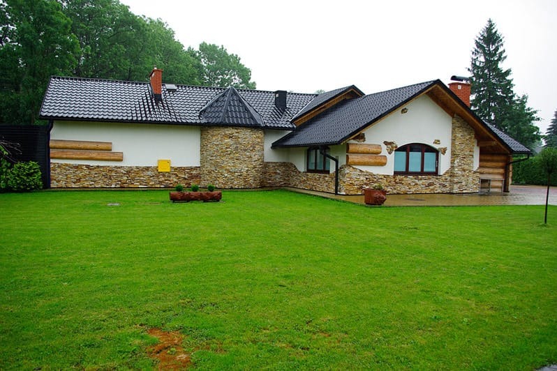 Czarny dach z dachówki ceramicznej na domu z elewacją z kamienia i drewna, otoczonym zielonym trawnikiem. Widok z przodu, pochmurny dzień.