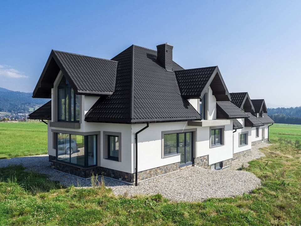 Nowoczesny dom jednorodzinny z ciemnym dachem z blachodachówki, jasną elewacją i dużymi oknami, otoczony zielonym terenem i górskim krajobrazem w tle.