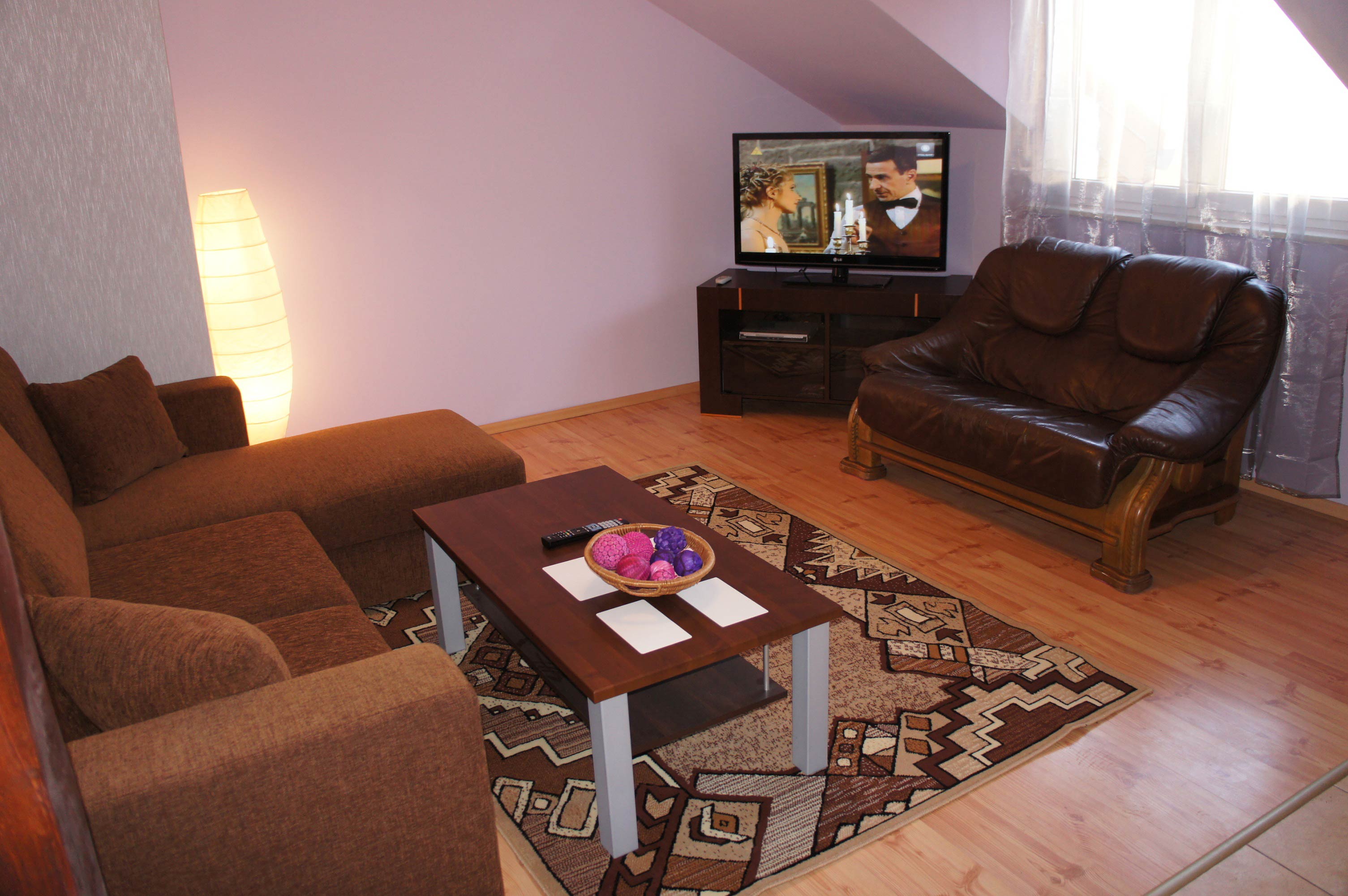 Jasny salon z brązowym narożnikiem, skórzaną sofą, telewizorem wyświetlającym scenę ślubną i stolikiem kawowym z dekoracyjnymi kulami w koszyku.