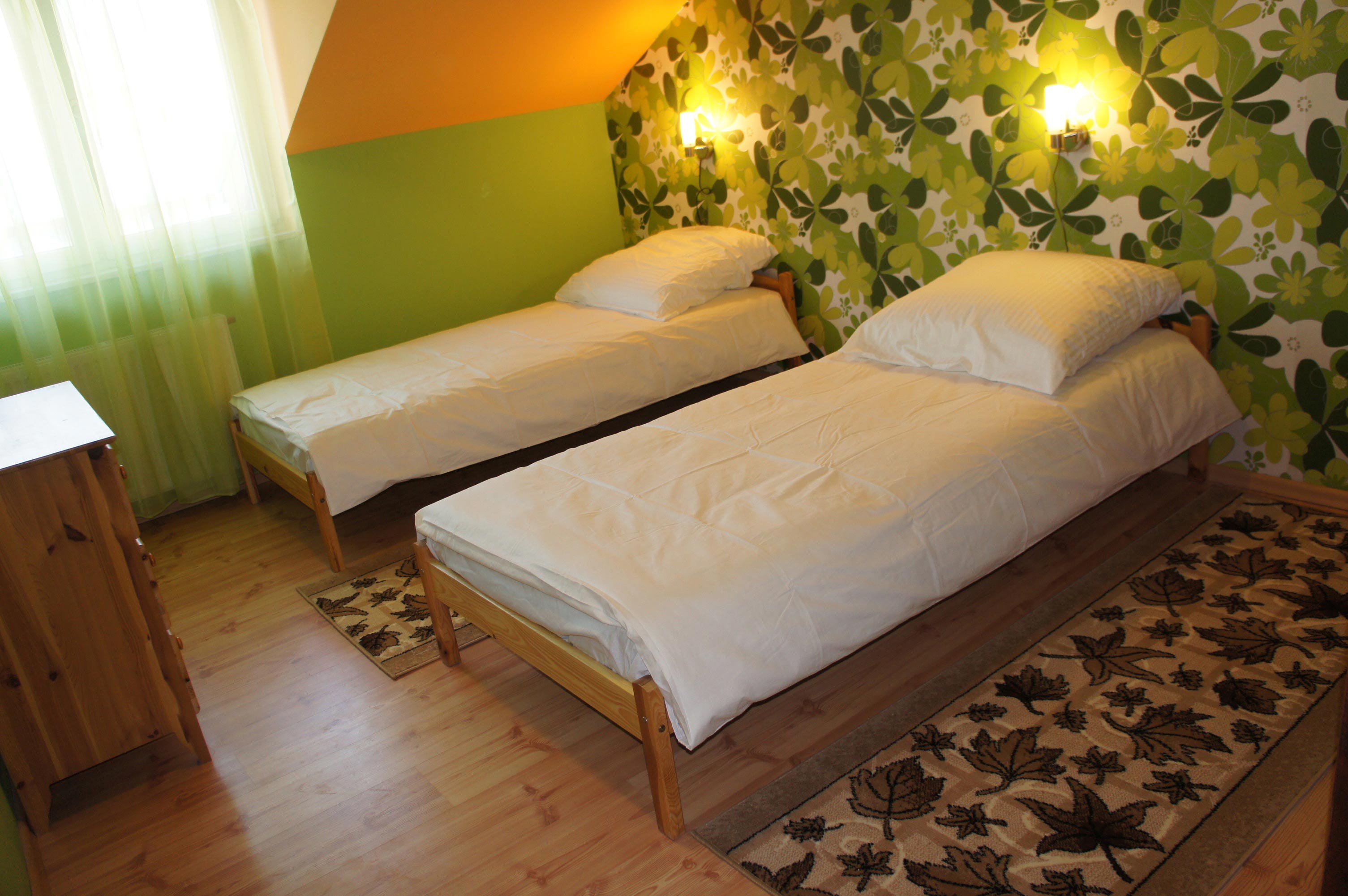 Pokój z dwoma łóżkami pojedynczymi w Rzeszowie, drewniane ramy, biała pościel, wzorzysta tapeta w kwiaty i dywan z motywem liści.