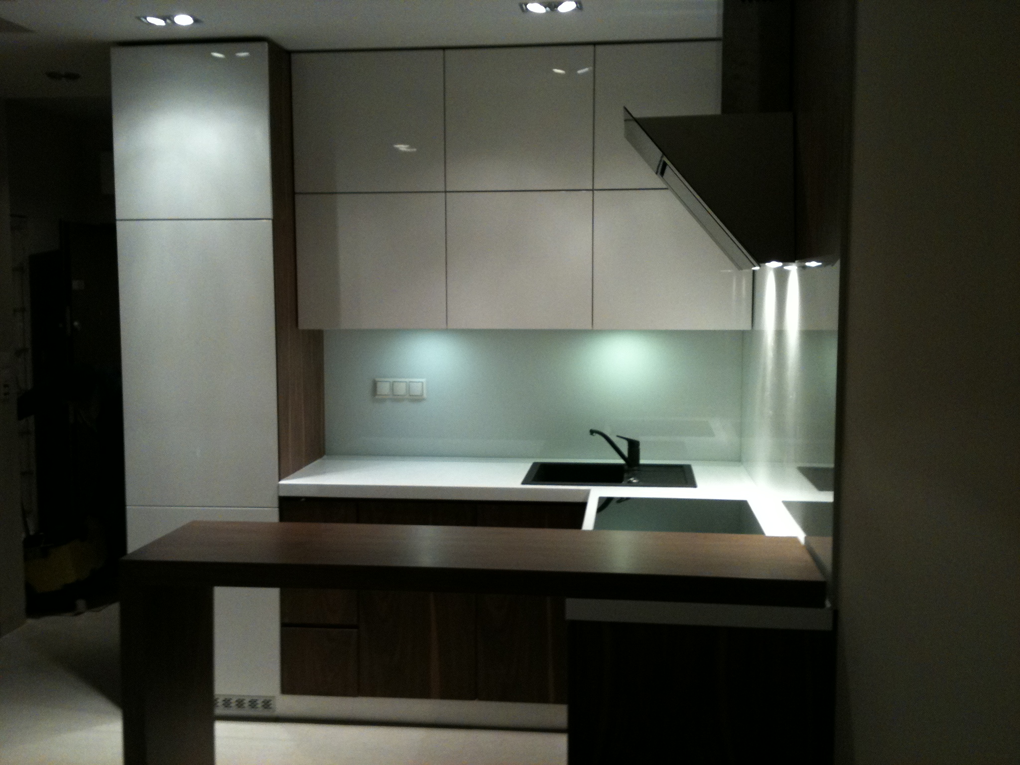 Nowoczesna kuchnia z białymi szafkami górnymi, drewnopodobnymi szafkami dolnymi i ciemnym blatem wyspowym, oświetlona punktowo, z czarnym zlewem i okapem.