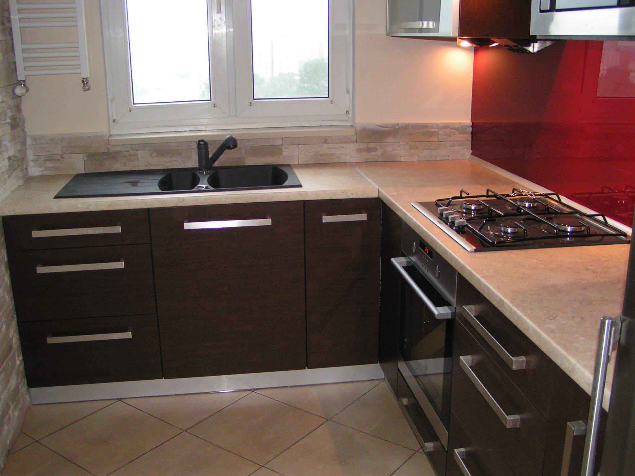 Kuchenna zabudowa narożna z ciemnobrązowymi frontami, beżowym blatem, płytą gazową i zlewozmywakiem, wykończona czerwoną szklaną ścianą i kamienną mozaiką nad blatem.
