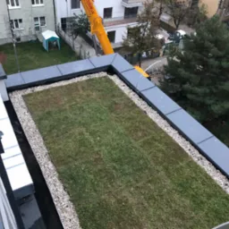 Zielony dach z obrzeżem z białych kamieni, widok z góry na budynek mieszkalny, w tle żółty dźwig i zabudowa osiedlowa.