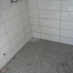 Wykończona kabina prysznicowa z szarymi, drobnymi kamieniami na podłodze i białymi, prostokątnymi płytkami na ścianach. Widoczny odpływ liniowy i metalowe uchwyty.