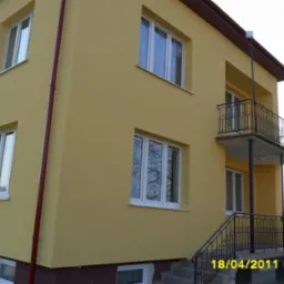 Żółty dom dwupiętrowy z balkonem i białymi oknami, widziany z dołu pod kątem. Widoczne schody wejściowe i stos rur oparty o balustradę.