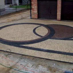 Ułożona kostka brukowa w kształcie dwóch przenikających się okręgów w kolorach beżu, brązu i czerni, widoczne garaże z cegły w tle, teren po budowie.