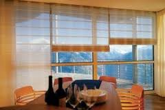 Jasne, poziome osłony okienne w ciepłym odcieniu, widoczne na tle rozległego krajobrazu górskiego przez duże okno w jadalni z zastawionym stołem.