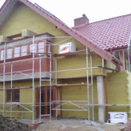 Dom w trakcie ocieplania, widoczne rusztowania, ceglany balkon i żółta izolacja. Czerwona dachówka i komin. Na ziemi cegły i ziemia.