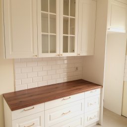 Gasiński Piotr - Jasna kuchnia z drewnianym blatem i białymi szafkami z przeszklonymi frontami. Widoczne złote uchwyty i białe płytki na ścianie. Klasyczny styl.