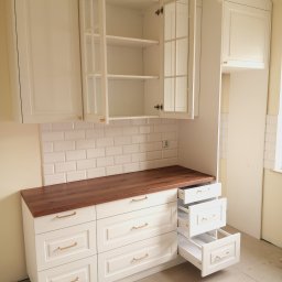 Gasiński Piotr - Biała zabudowa kuchenna z drewnianym blatem i otwartymi szufladami, płytki metro na ścianie, oświetlenie sufitowe. Fronty szafek frezowane, uchwyty złote.