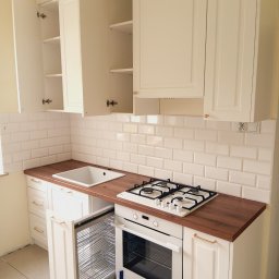 Gasiński Piotr - Biała kuchnia z drewnianym blatem. Otwarta szafka z koszem cargo, białe płytki na ścianie. U góry szafki otwarte.