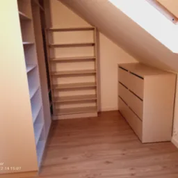 Jasna zabudowa wnęki na poddaszu: szafa z półkami, komoda z szufladami, drewnopodobna podłoga, okno dachowe. Całość w neutralnych barwach.