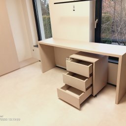 Gasiński Piotr - Beżowe biurko na wymiar z trzema wysuniętymi szufladami, ustawione przy oknie z widokiem na zieleń. Minimalistyczny design, jasne wnętrze, widoczne gniazdko elektryczne.