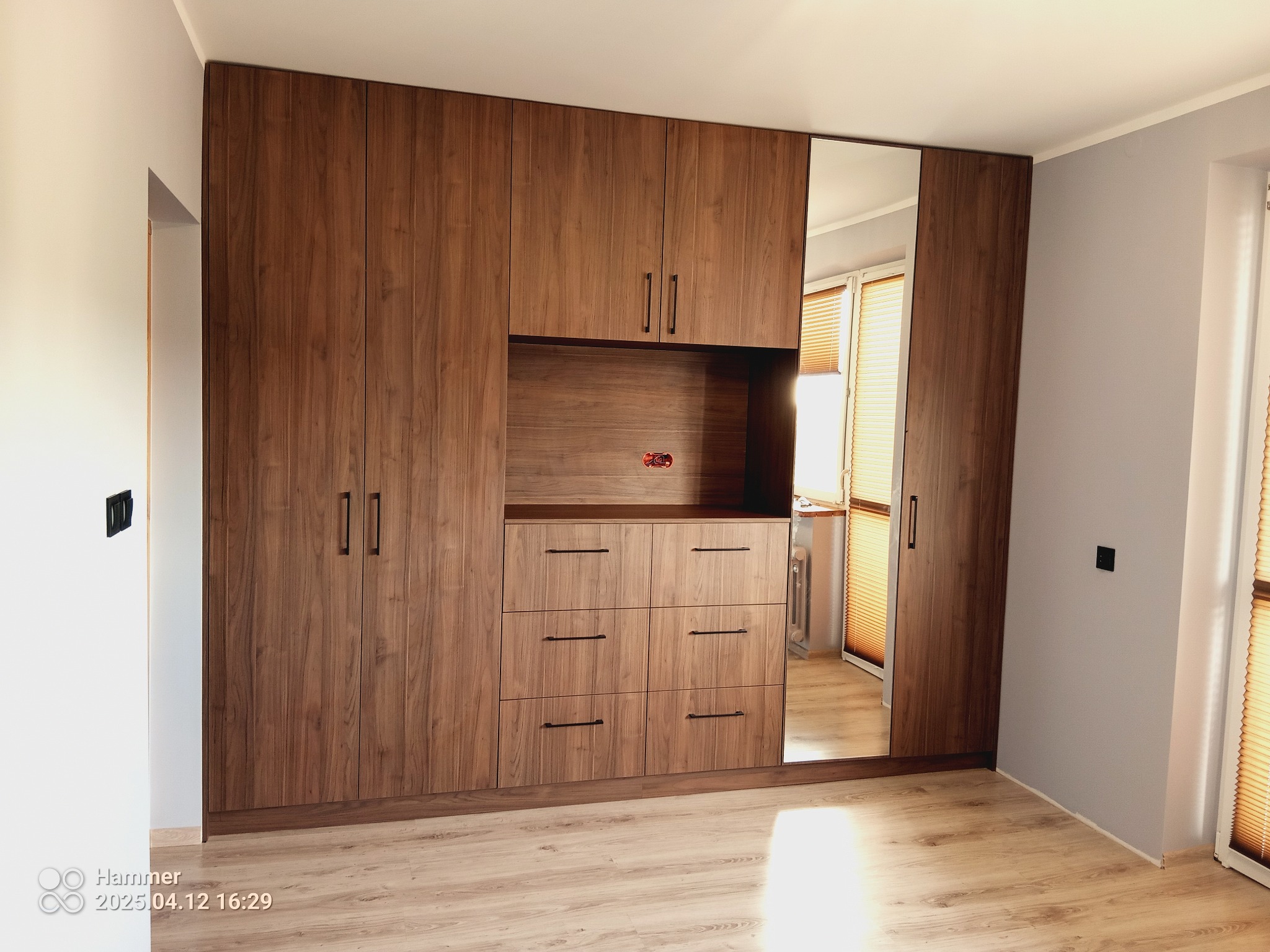 Duża, drewniana szafa wnękowa z lustrem, wkomponowana w jasne wnętrze. Widoczne szuflady i półki, minimalistyczne uchwyty. Całość utrzymana w ciepłej tonacji.