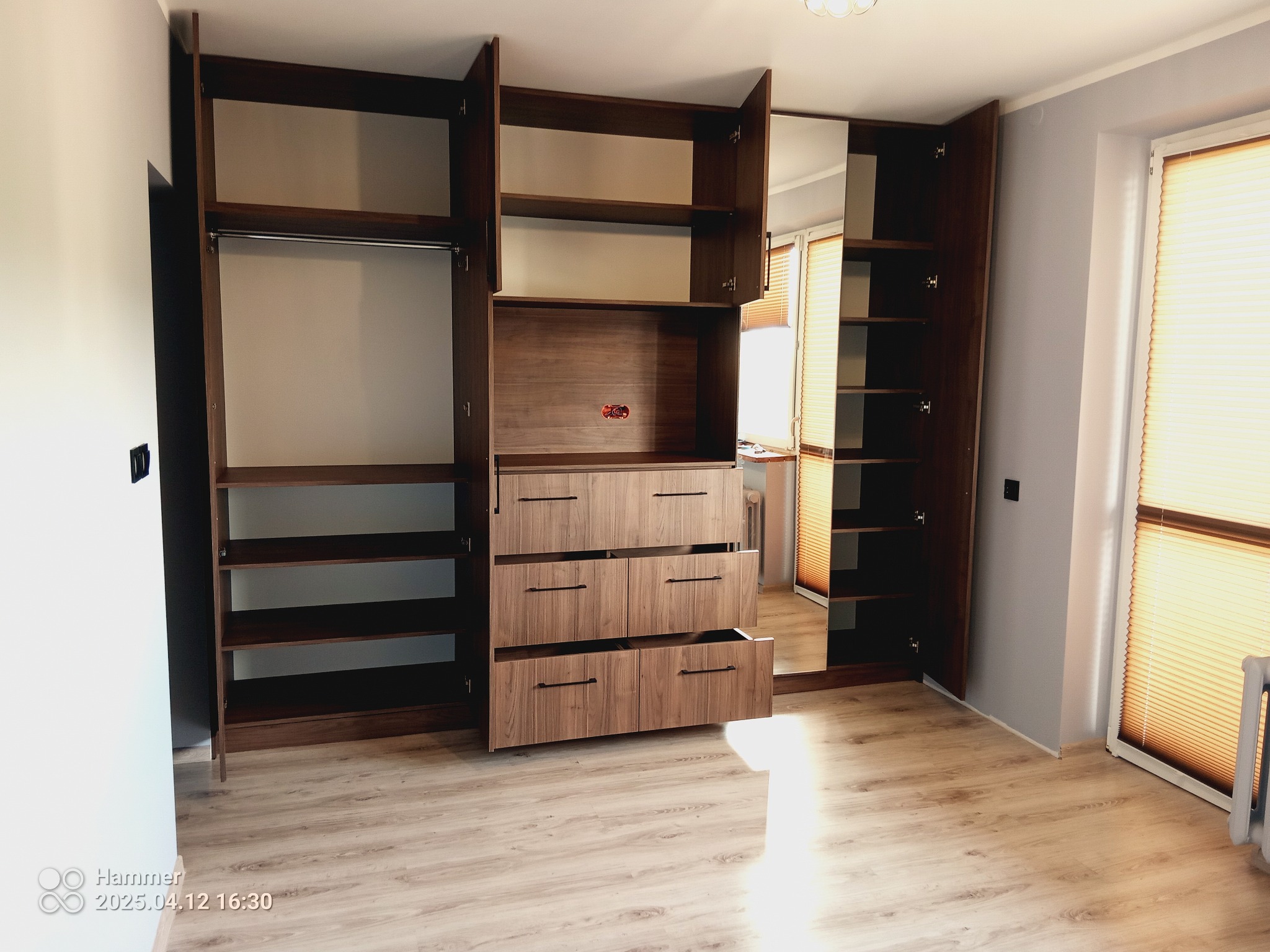 Przestronna szafa wnękowa z drewnianymi frontami i lustrem, częściowo otwarta, ukazująca półki i drążek na ubrania. Jasne wnętrze, widok na okno w odbiciu.