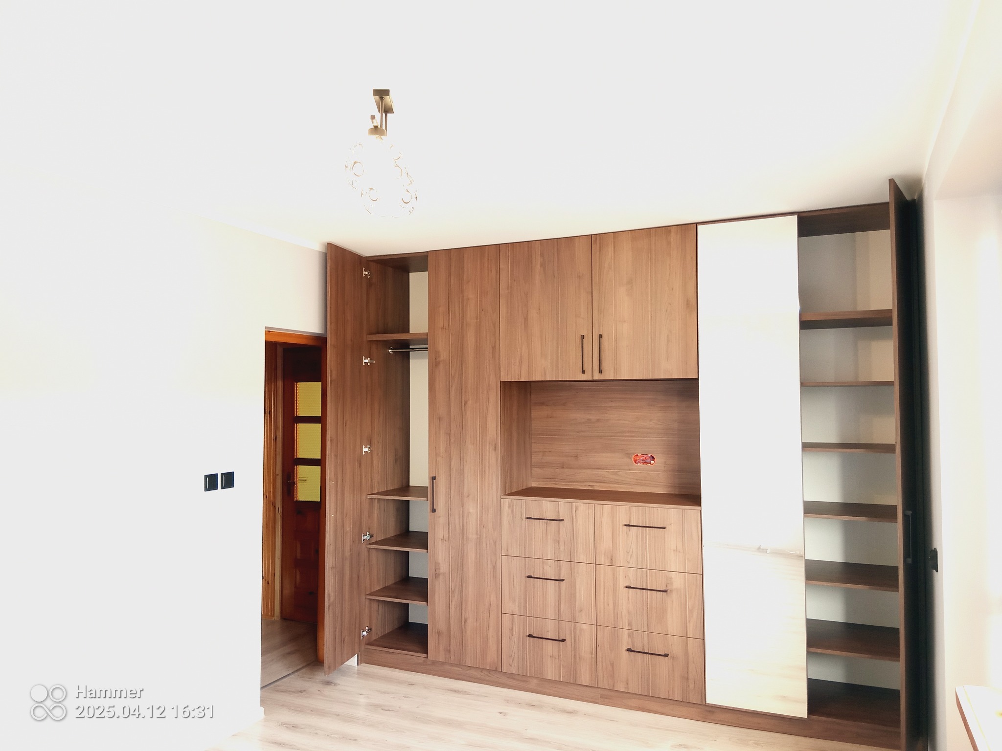 Drewniana szafa wnękowa z otwartymi drzwiami, półkami i szufladami, uzupełniona białym segmentem. Widoczne gniazdko elektryczne w środkowej części szafy.