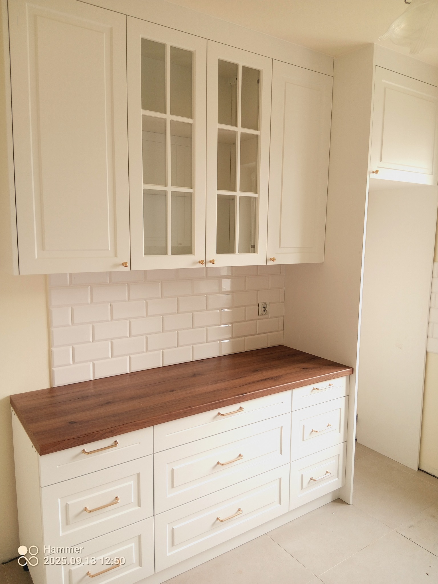 Jasna kuchnia z drewnianym blatem i białymi szafkami z przeszklonymi frontami. Widoczne złote uchwyty i białe płytki na ścianie. Klasyczny styl.