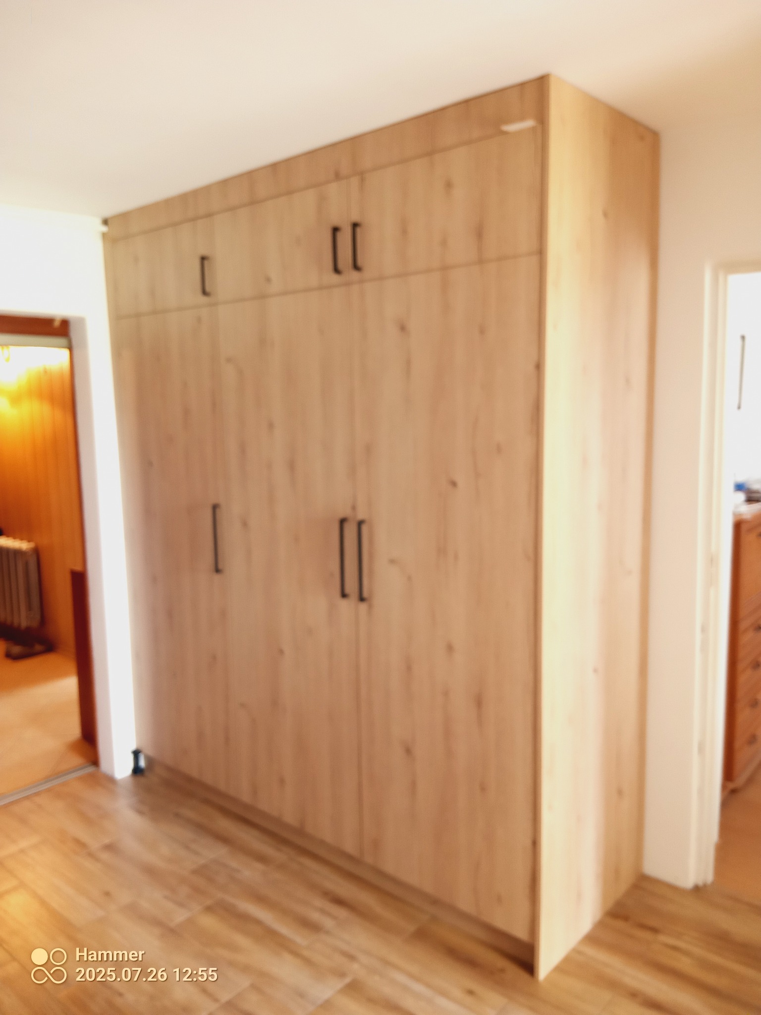 Drewniana szafa wnękowa z ciemnymi uchwytami, dopasowana do podłogi, z widocznymi drzwiami do innego pomieszczenia.