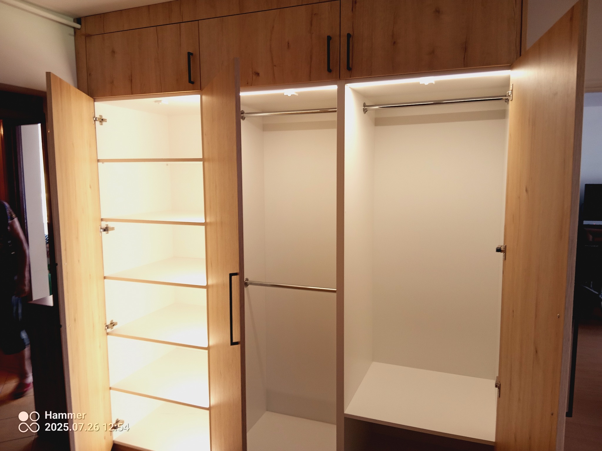 Jasna szafa wnękowa z oświetleniem LED, otwartymi drzwiami ukazującymi półki i drążki na ubrania. Widoczne czarne, podłużne uchwyty.