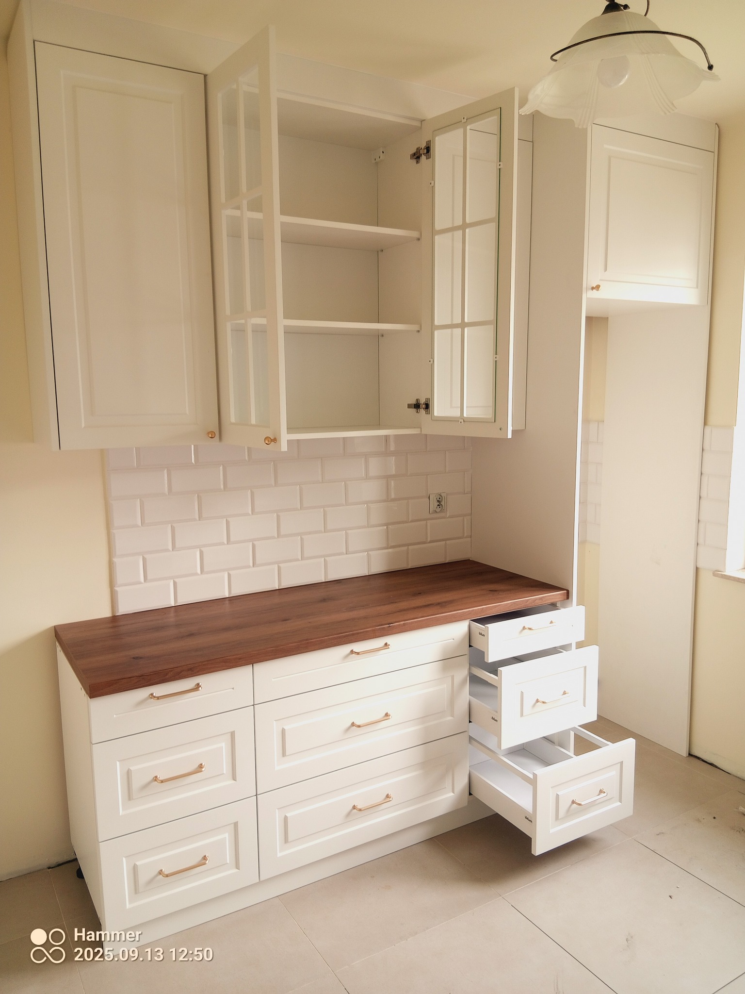 Biała zabudowa kuchenna z drewnianym blatem i otwartymi szufladami, płytki metro na ścianie, oświetlenie sufitowe. Fronty szafek frezowane, uchwyty złote.