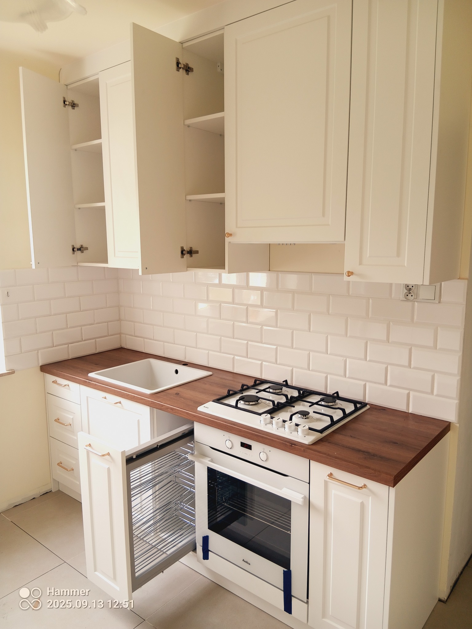 Biała kuchnia z drewnianym blatem. Otwarta szafka z koszem cargo, białe płytki na ścianie. U góry szafki otwarte.