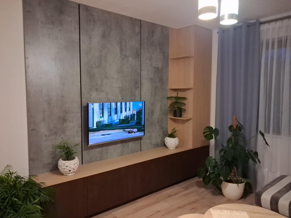 Nowoczesna ściana TV z szarą okładziną imitującą beton, drewnianymi szafkami i półkami. Telewizor zawieszony na ścianie, rośliny doniczkowe dodają przytulności.