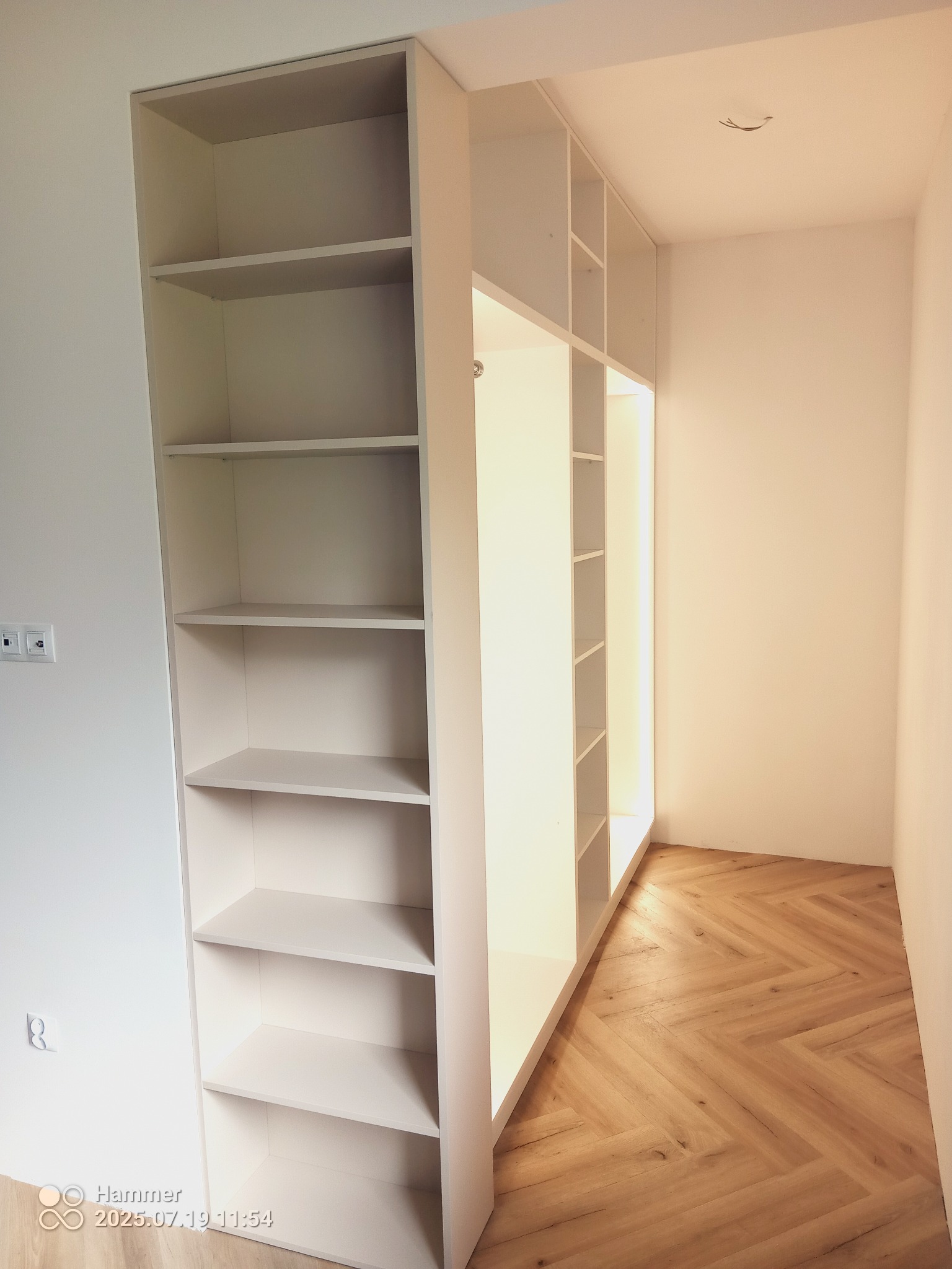 Biała, modułowa szafa z półkami wnękowymi, oświetlona od wewnątrz, na drewnianej podłodze ułożonej w jodełkę, w minimalistycznym wnętrzu.