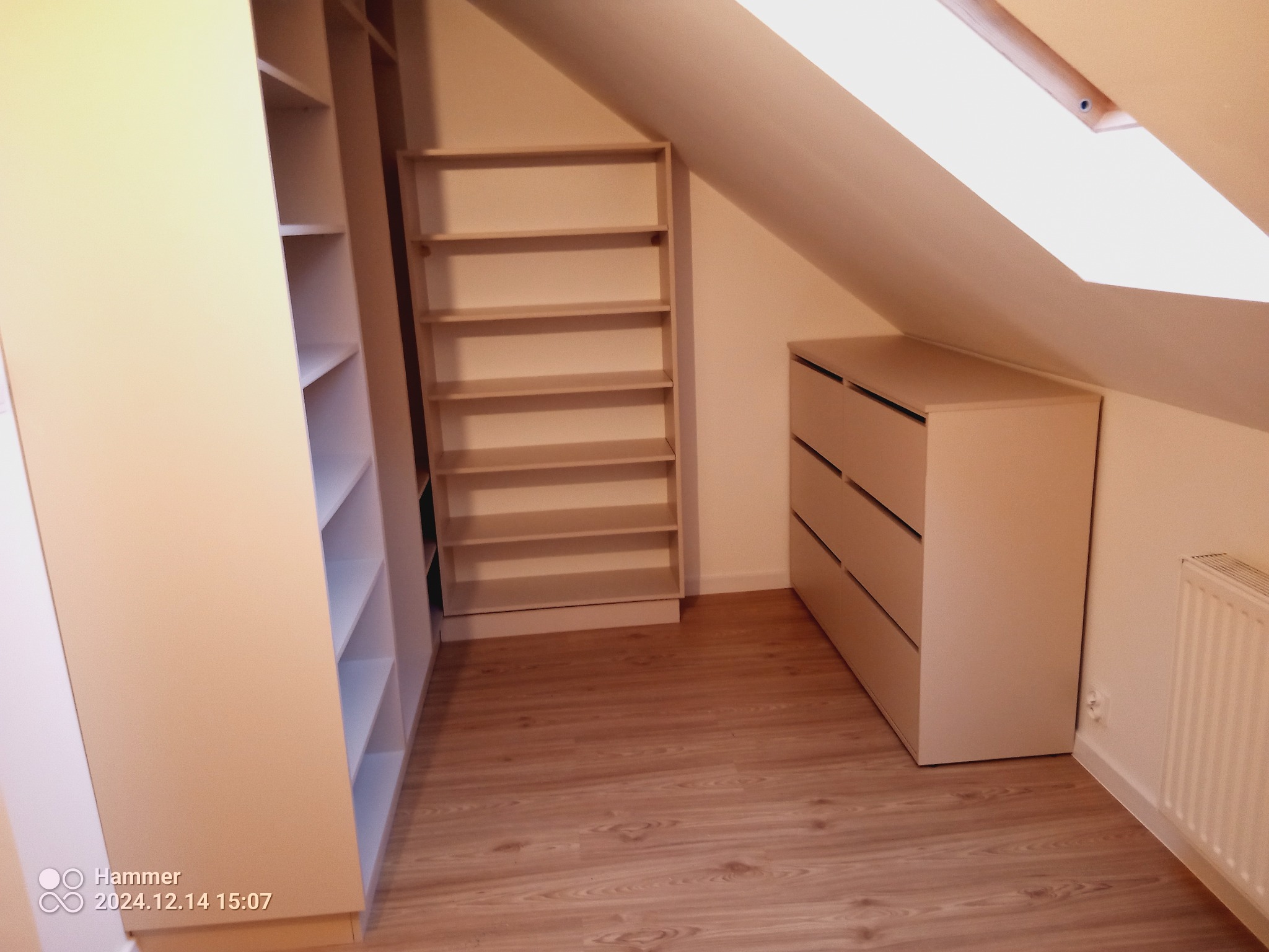 Jasna zabudowa wnęki na poddaszu: szafa z półkami, komoda z szufladami, drewnopodobna podłoga, okno dachowe. Całość w neutralnych barwach.